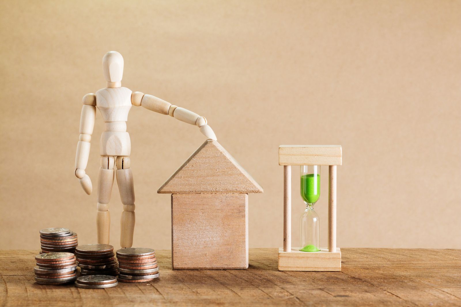 Holzhaus und Gliederfigur neben Münzstapeln und einer Sanduhr als Symbol für Immobilienplanung.